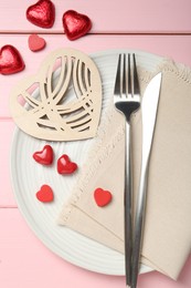 Photo of Romantic place setting for Valentine's day. Cutlery, napkin, plate and decorative hearts on pink wooden table, flat lay