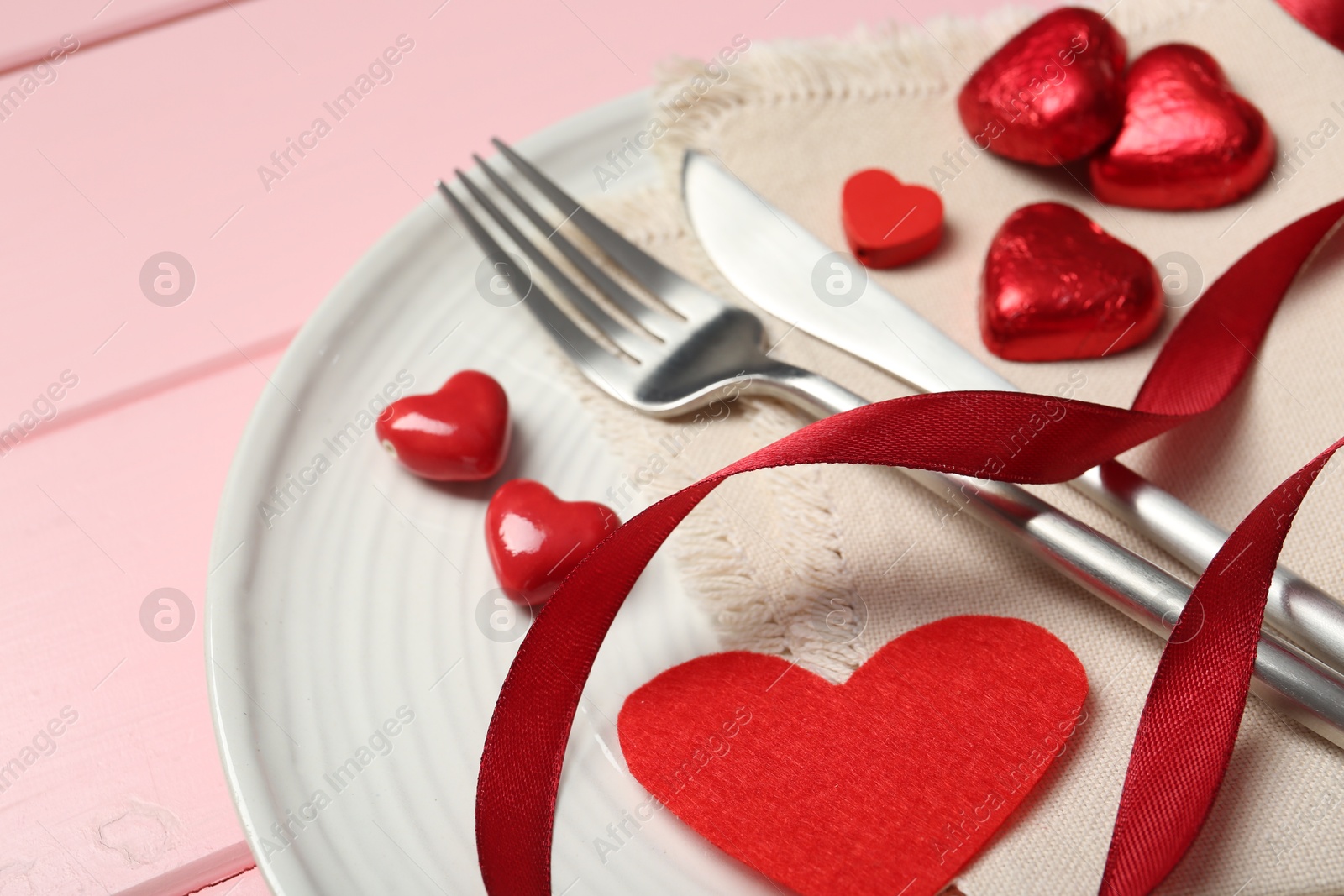 Photo of Romantic place setting for Valentine's day. Cutlery, napkin, plate and decorative hearts on pink wooden table, closeup