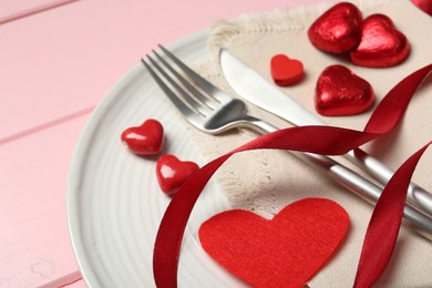 Photo of Romantic place setting for Valentine's day. Cutlery, napkin, plate and decorative hearts on pink wooden table, closeup