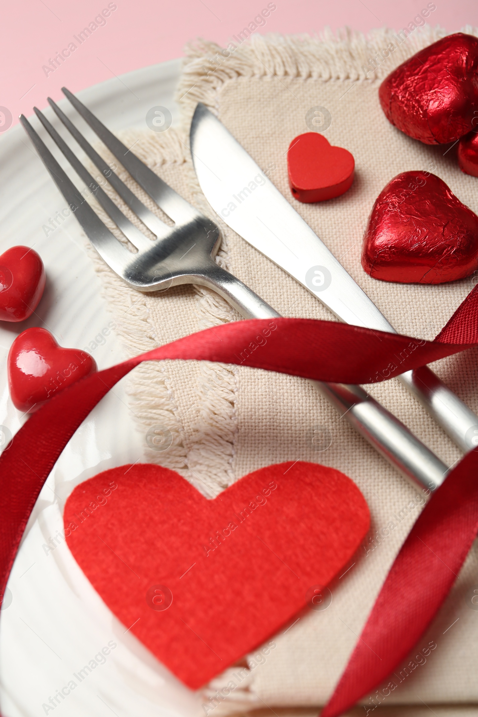 Photo of Romantic place setting for Valentine's day. Cutlery, napkin, plate and decorative hearts on pink wooden table, closeup