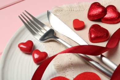 Photo of Romantic place setting for Valentine's day. Cutlery, napkin, plate and decorative hearts on pink wooden table, closeup