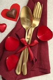 Romantic place setting for Valentine's day. Cutlery, napkin, candy, petals and decorative hearts on light textured table, flat lay