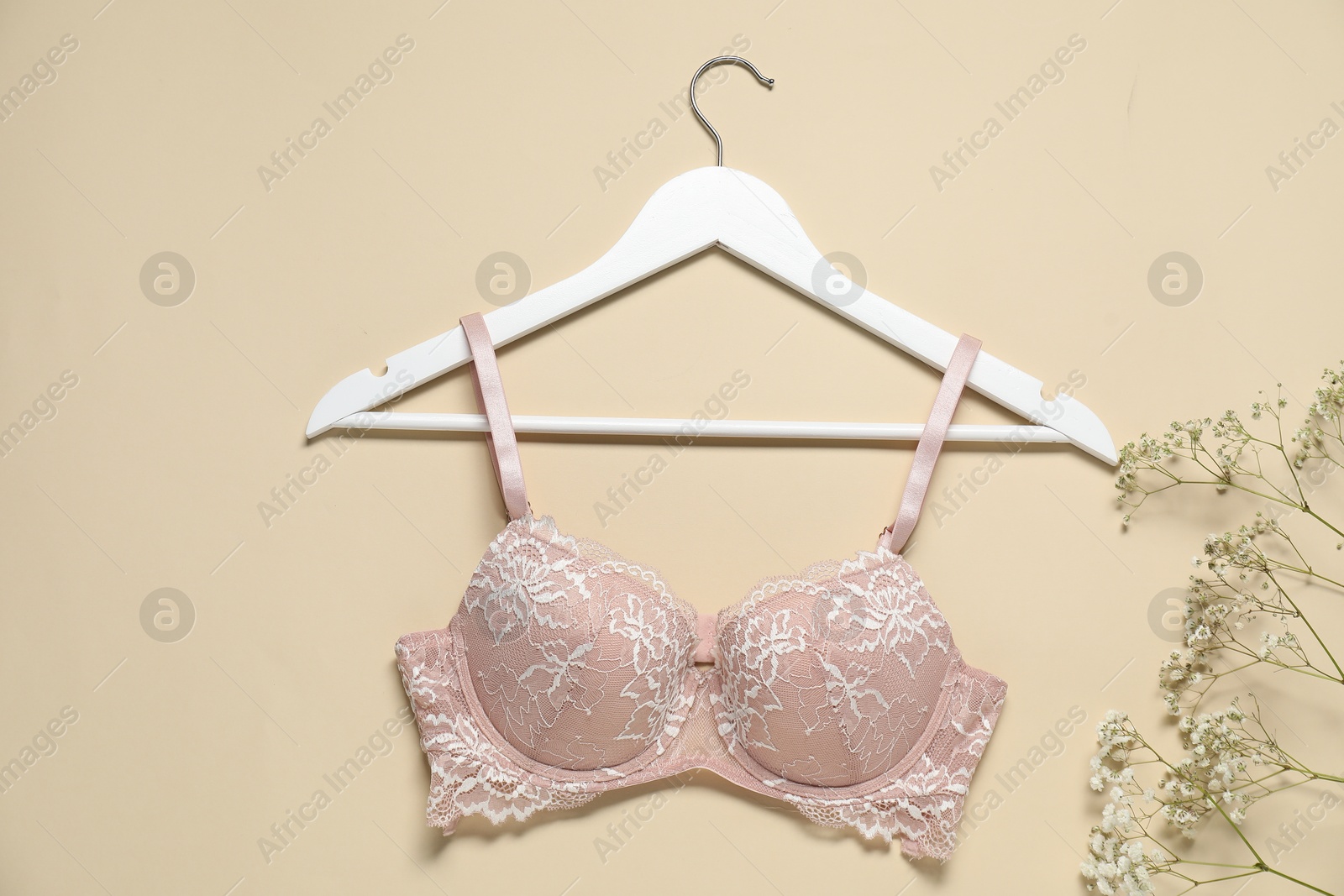 Photo of Hanger with beautiful lace bra and flowers on beige background, flat lay