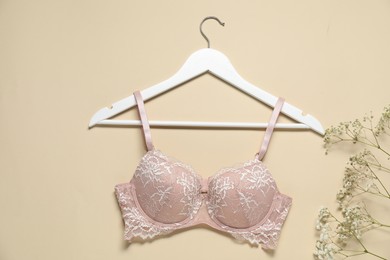 Photo of Hanger with beautiful lace bra and flowers on beige background, flat lay