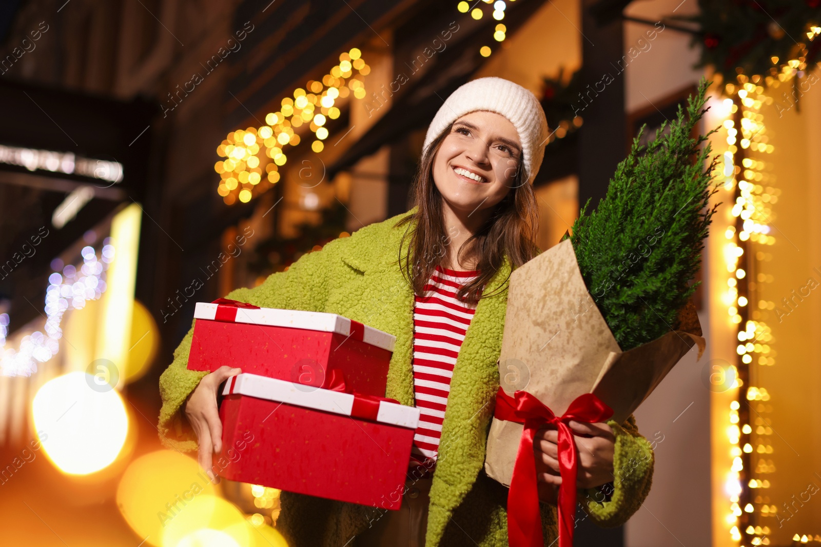 Photo of Happy woman with thuja tree and Christmas gifts outdoors, low angle view. Bokeh effect