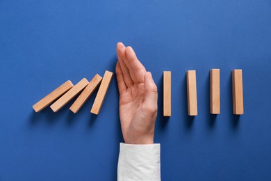 Photo of Man stopping wooden blocks from falling on blue background, top view. Domino effect