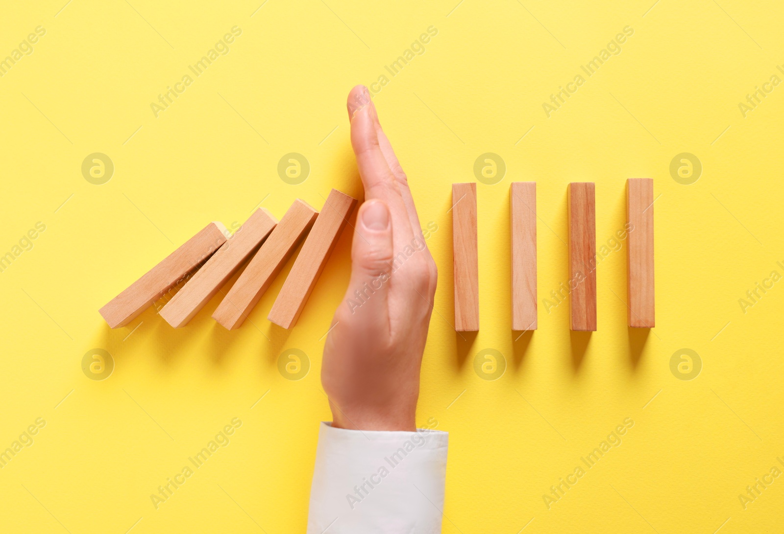 Photo of Man stopping wooden blocks from falling on yellow background, top view. Domino effect