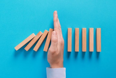 Photo of Man stopping wooden blocks from falling on light blue background, top view. Domino effect