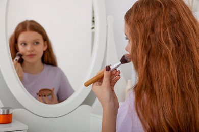 Photo of Teenage girl applying blush with brush near mirror indoors