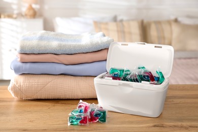 Photo of Detergent capsules in container and clean laundry on wooden table indoors