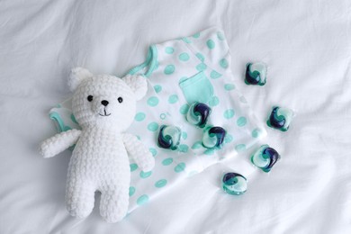 Photo of Laundry detergent capsules, teddy bear and baby onesie on white fabric, flat lay