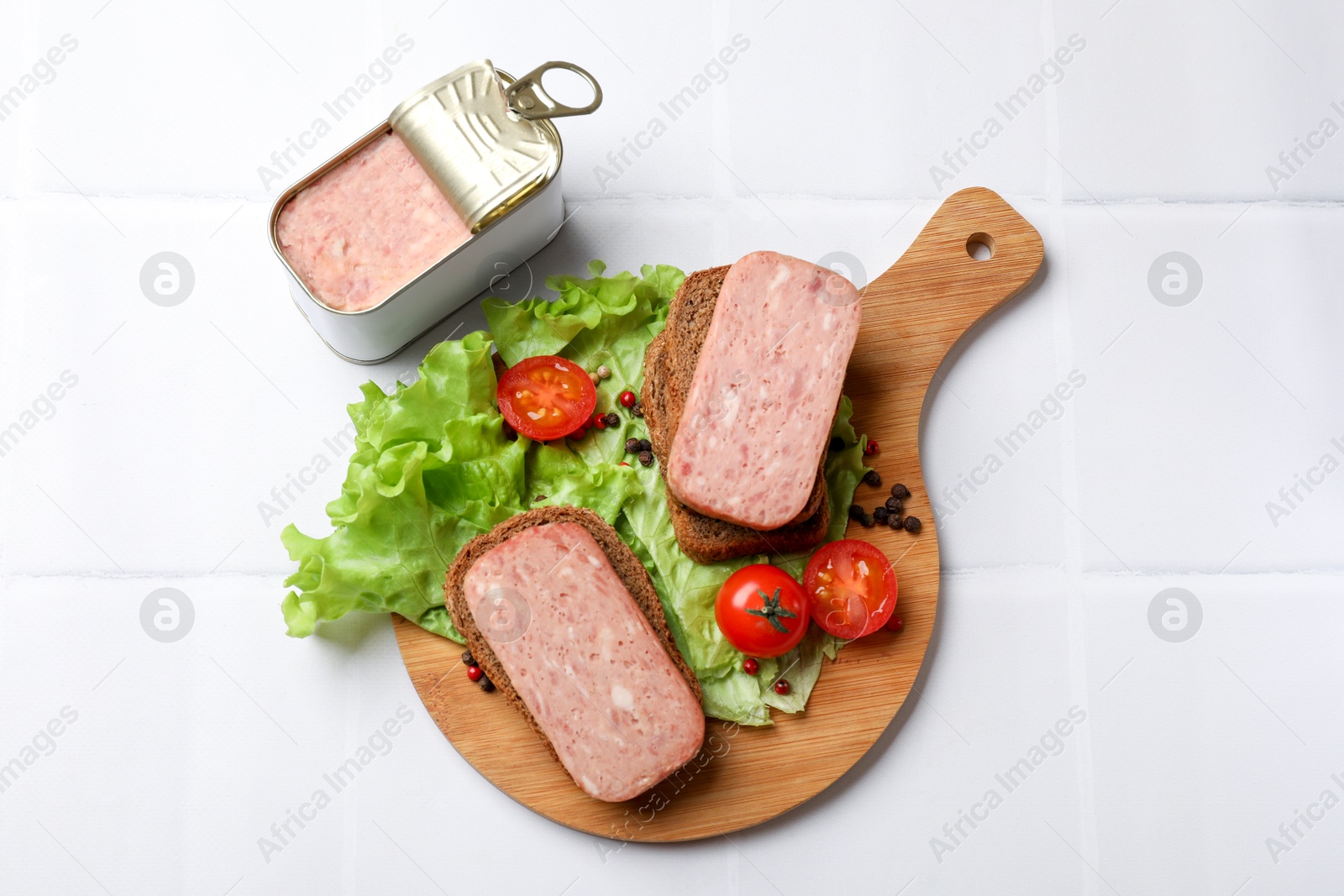 Photo of Tasty canned meat, bread, tomatoes, spices and lettuce on white tiled table, top view