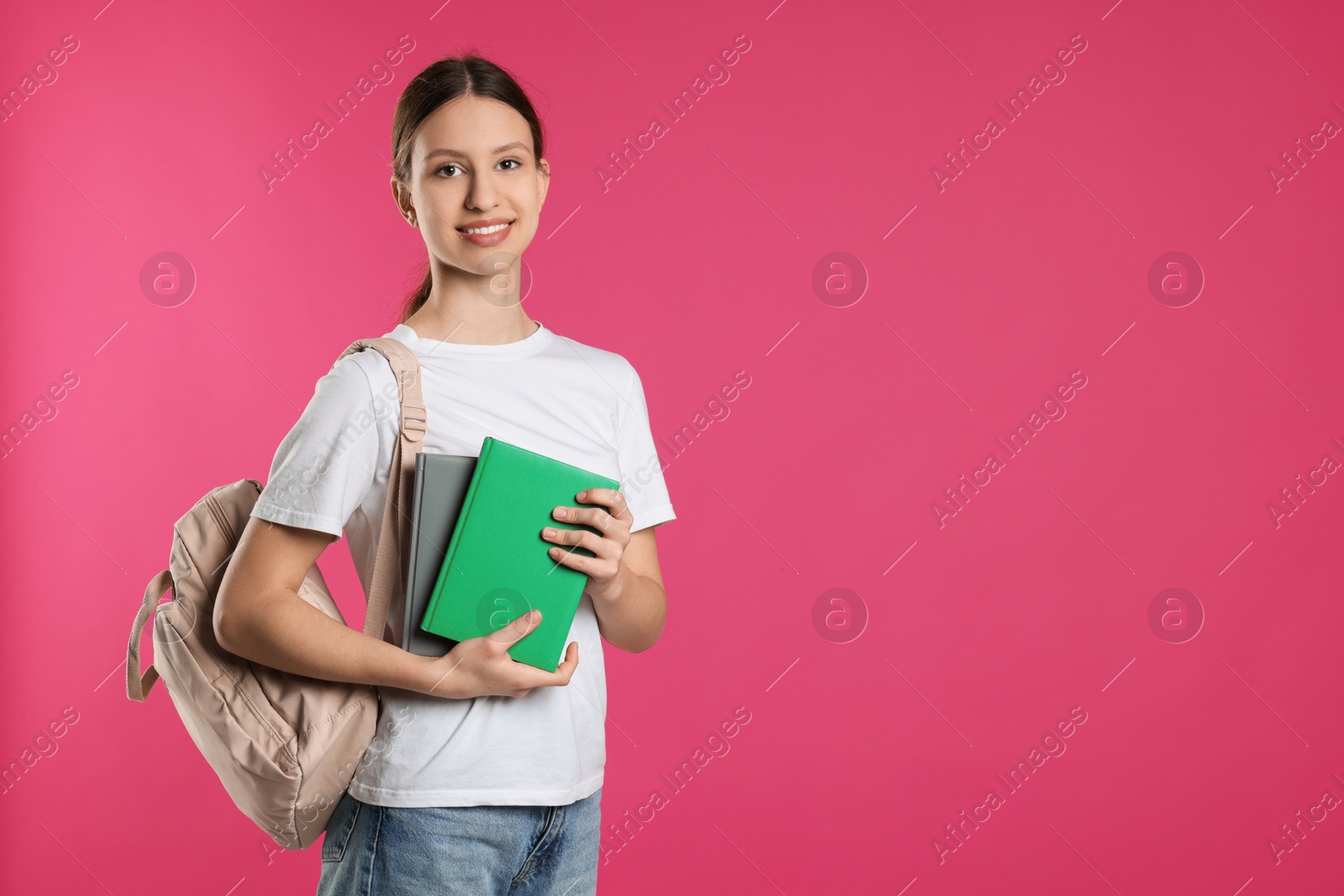 Photo of Portrait of smiling teenage girl with books and backpack on pink background. Space for text