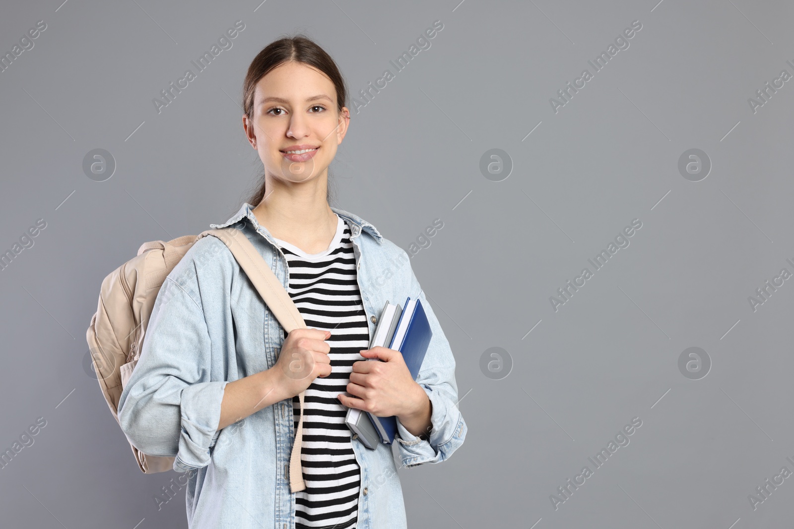 Photo of Portrait of smiling teenage girl with books and backpack on grey background. Space for text