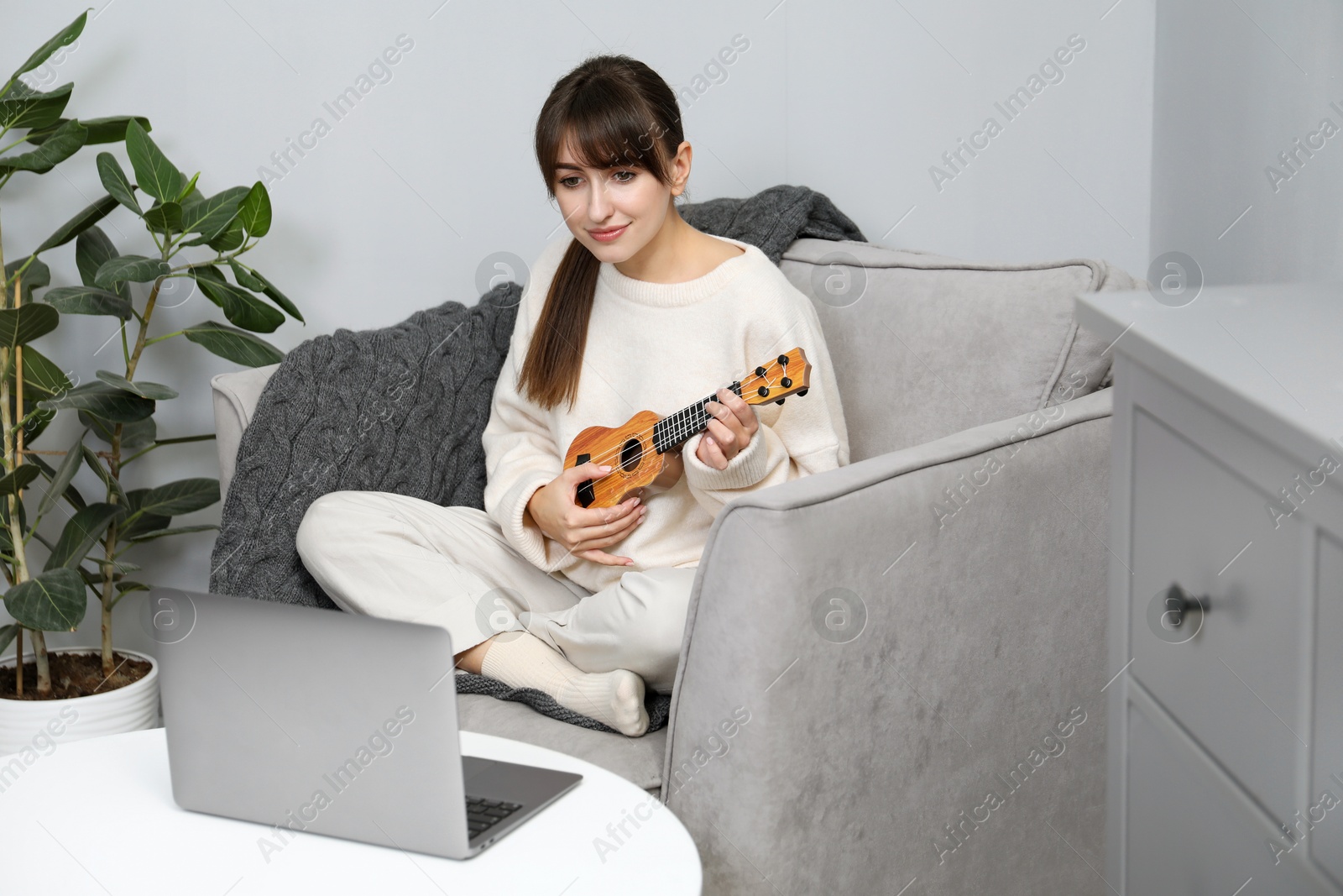 Photo of Woman learning to play ukulele with online music course in armchair at home