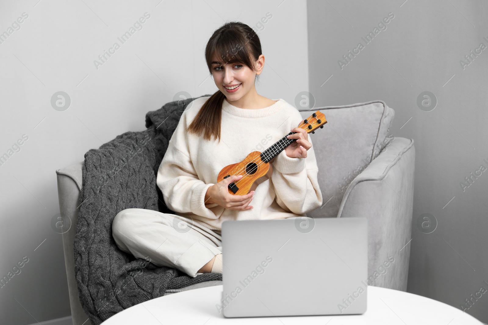 Photo of Woman learning to play ukulele with online music course in armchair at home