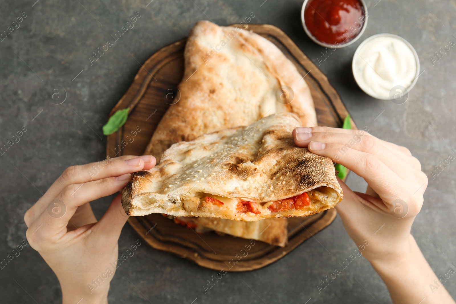 Photo of Woman eating tasty calzone with meat, cheese and tomato at grey textured table, top view