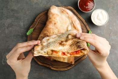 Photo of Woman eating tasty calzone with meat, cheese and tomato at grey textured table, top view