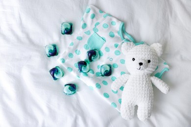 Photo of Laundry detergent capsules, teddy bear and baby onesie on white fabric, flat lay