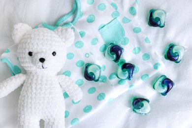 Photo of Laundry detergent capsules, teddy bear and baby onesie on white fabric, flat lay