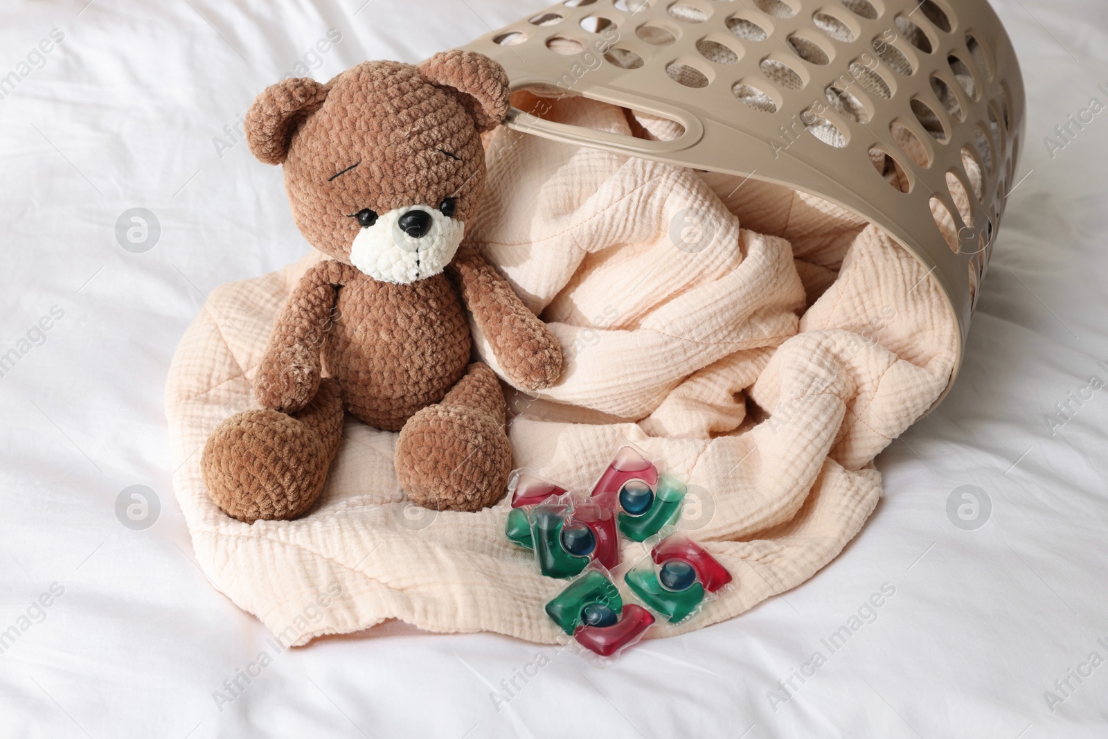 Photo of Laundry detergent capsules, teddy bear and basket with clean linens on bed