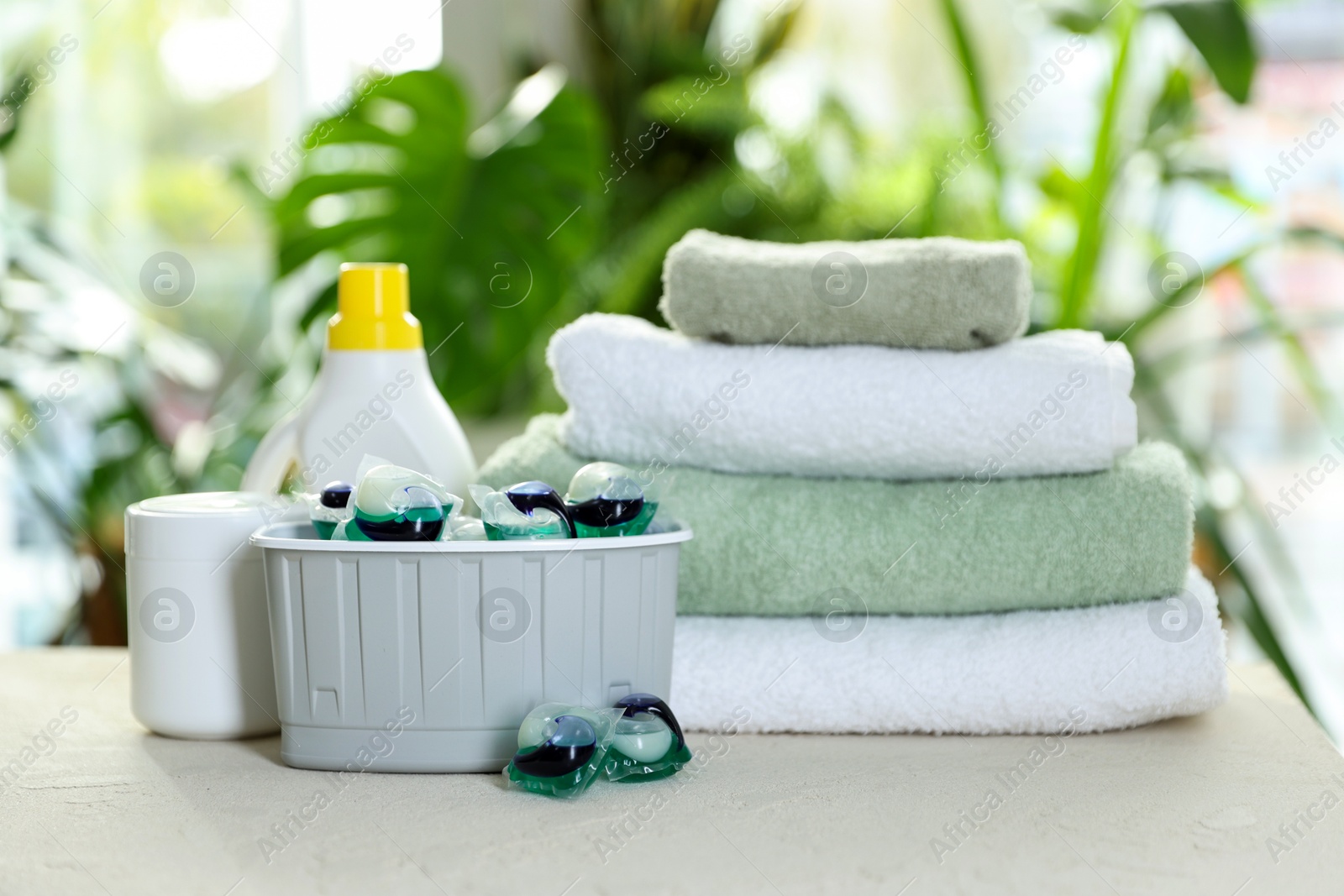 Photo of Container with laundry capsules, detergents and clean towels on table against blurred background, selective focus