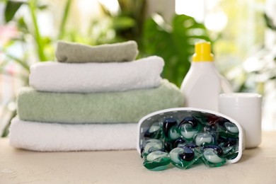 Photo of Container with laundry capsules, detergents and clean towels on table against blurred background, selective focus