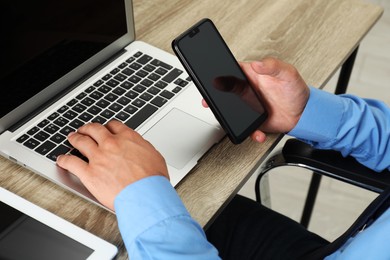 Photo of Businessman with smartphone using laptop at wooden table, closeup. Modern technology