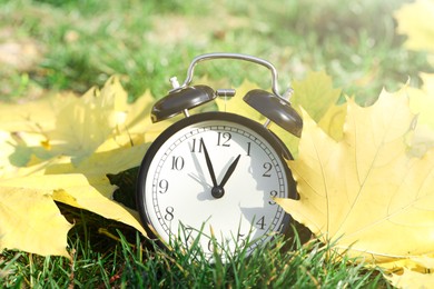 Photo of Autumn time. Alarm clock among fallen leaves on green grass, closeup