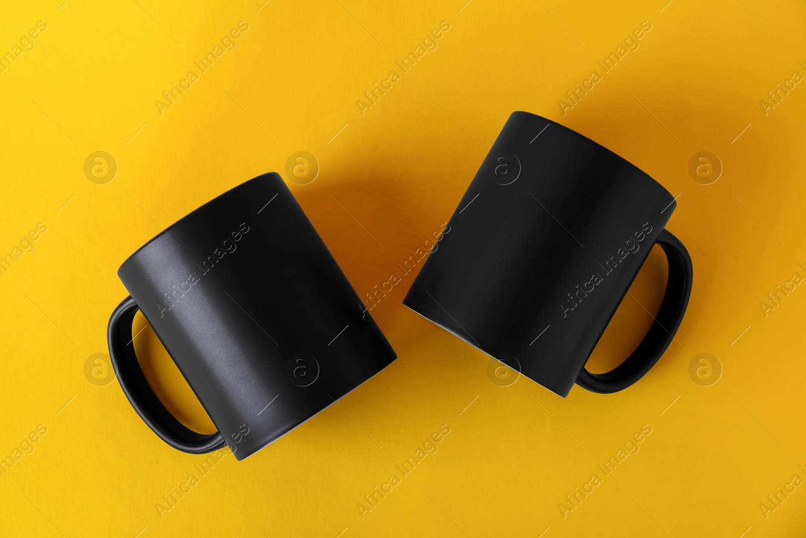 Photo of Two blank black mugs on yellow background, top view. Mockup for design