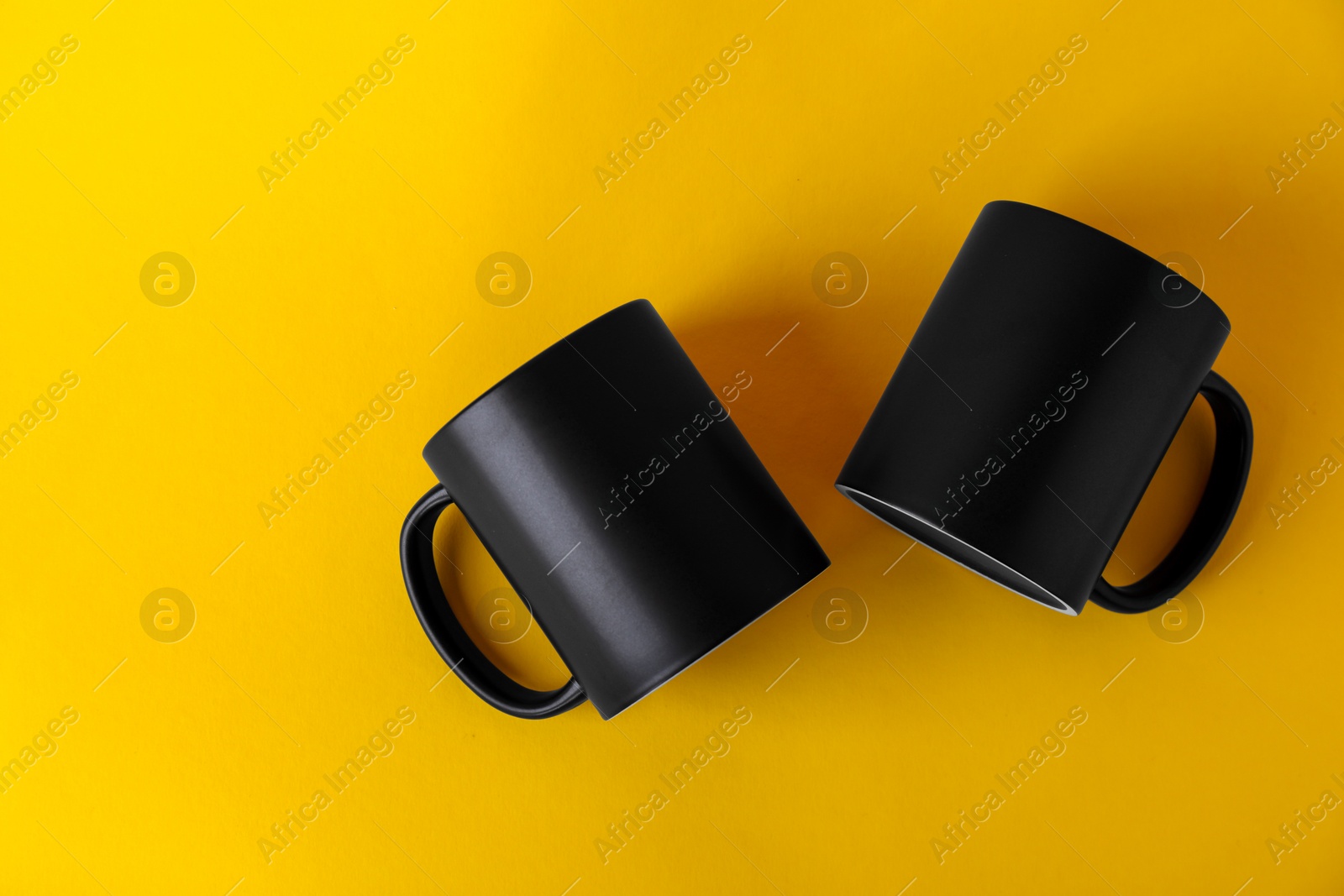 Photo of Two blank black mugs on yellow background, top view. Mockup for design