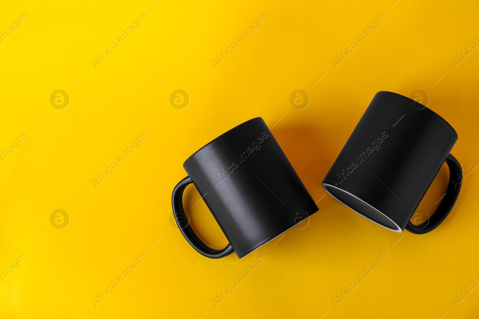 Photo of Two blank black mugs on yellow background, top view. Mockup for design