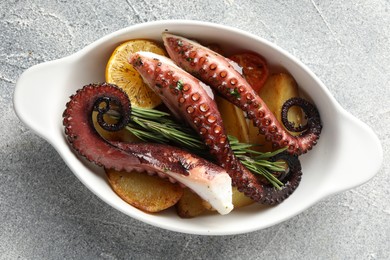 Photo of Fried octopus with vegetables, lemon and rosemary in baking dish on grey textured table, top view