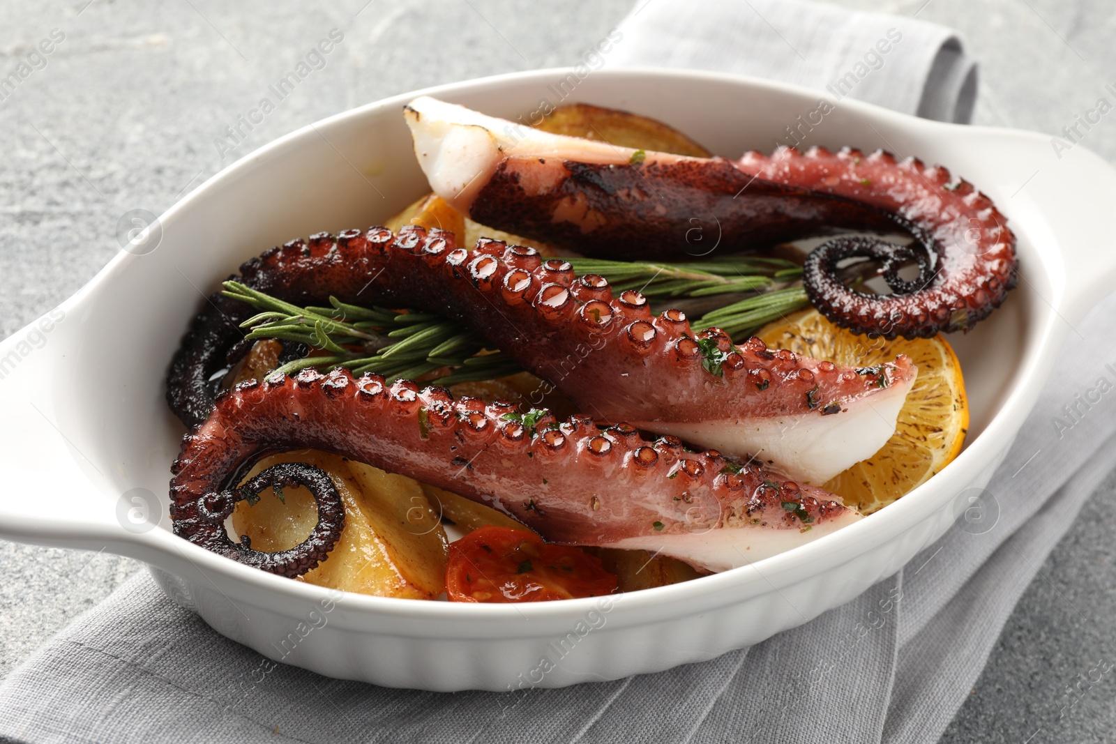 Photo of Fried octopus with vegetables, lemon and rosemary in baking dish on grey textured table, closeup