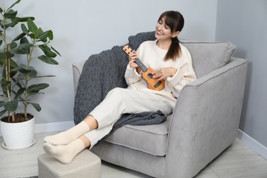 Happy woman playing ukulele in armchair at home