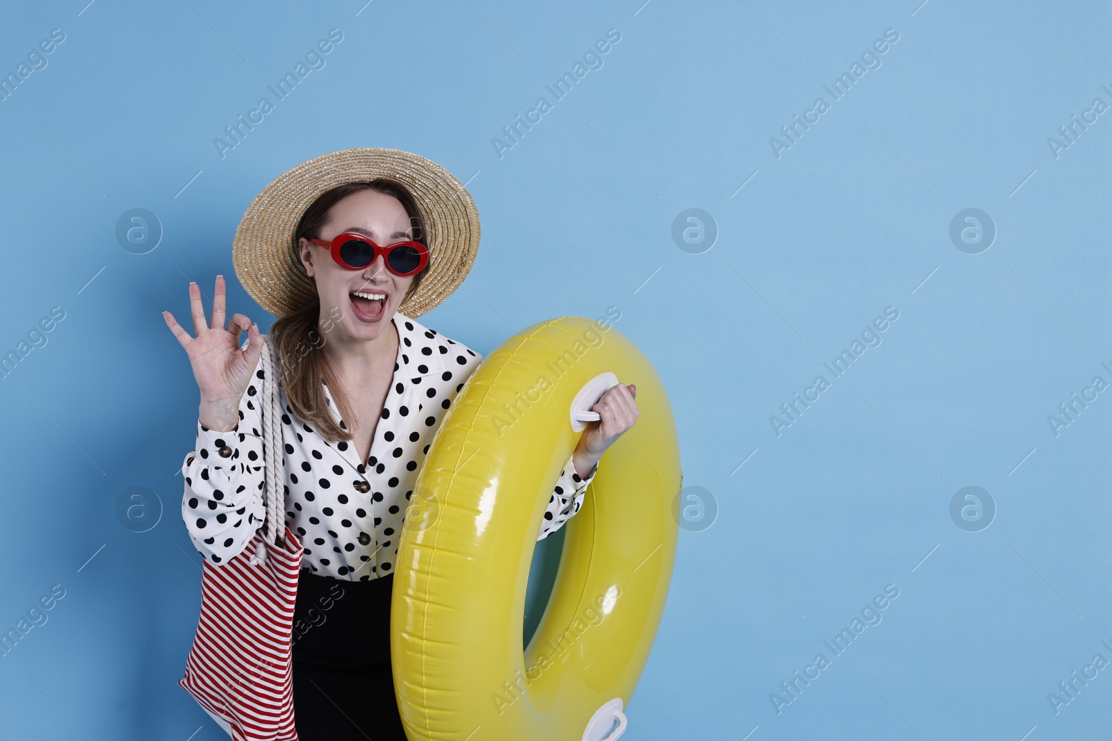 Photo of Businesswoman with inflatable ring, straw hat, bag and sunglasses showing ok gesture on light blue background, space for text