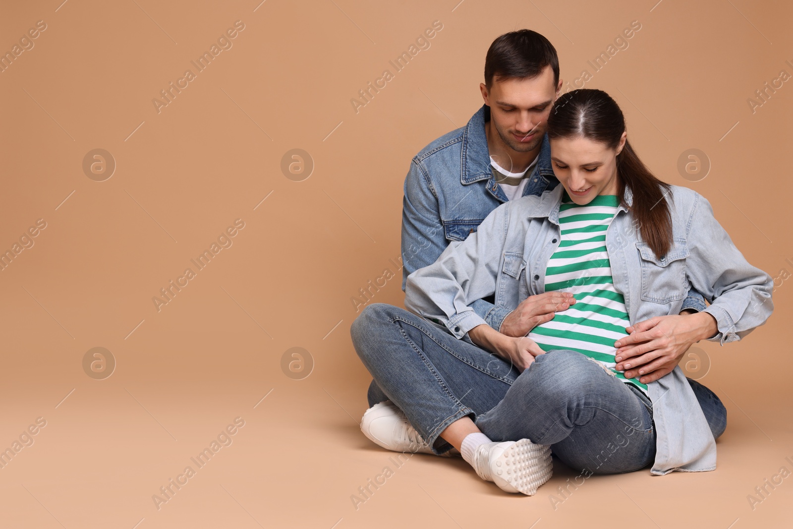 Photo of Man trying to feel baby kicks in his pregnant wife's belly on beige background. Space for text