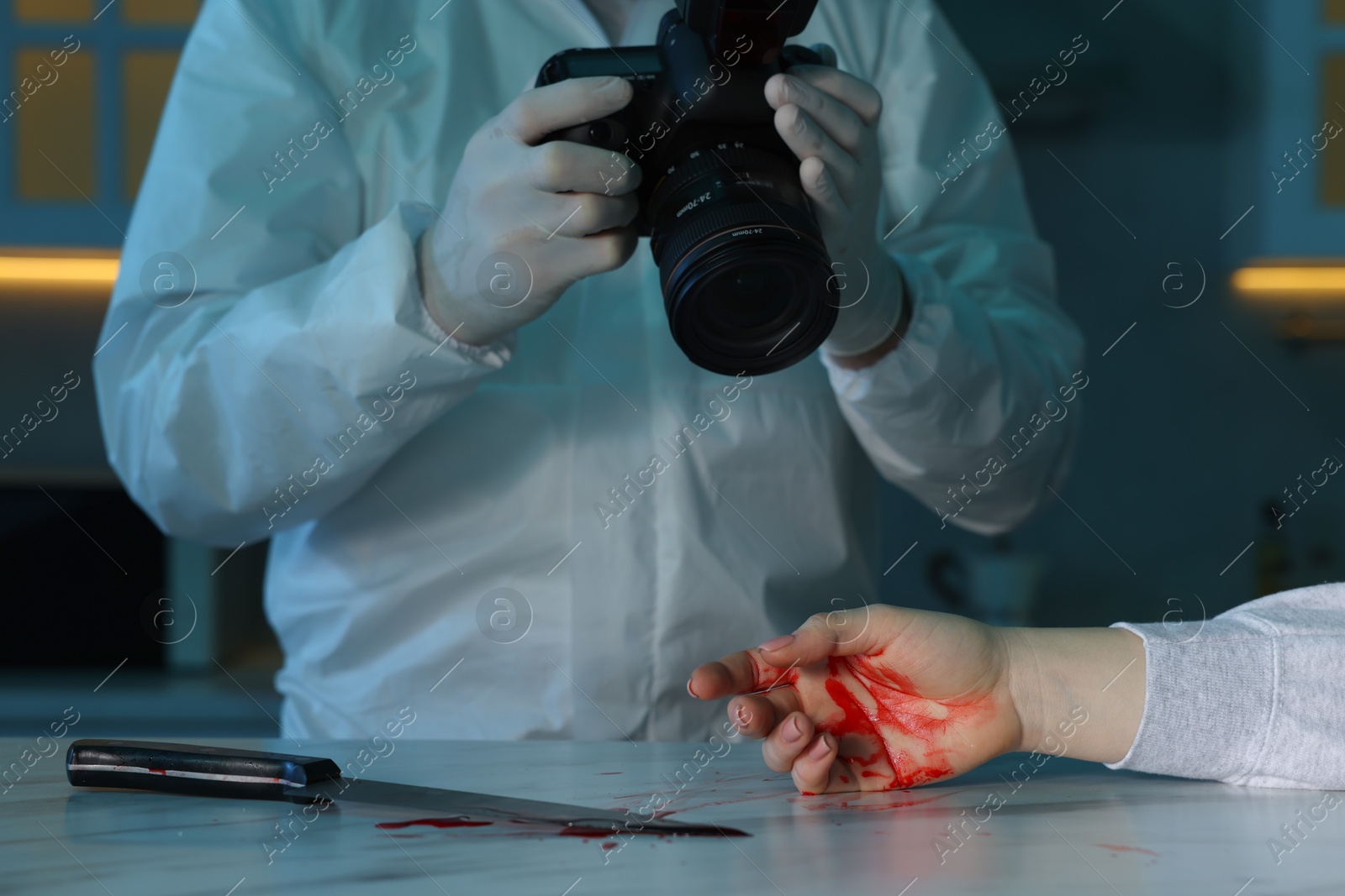 Photo of Forensic expert with camera, knife, blood and dead woman's body at countertop indoors, closeup