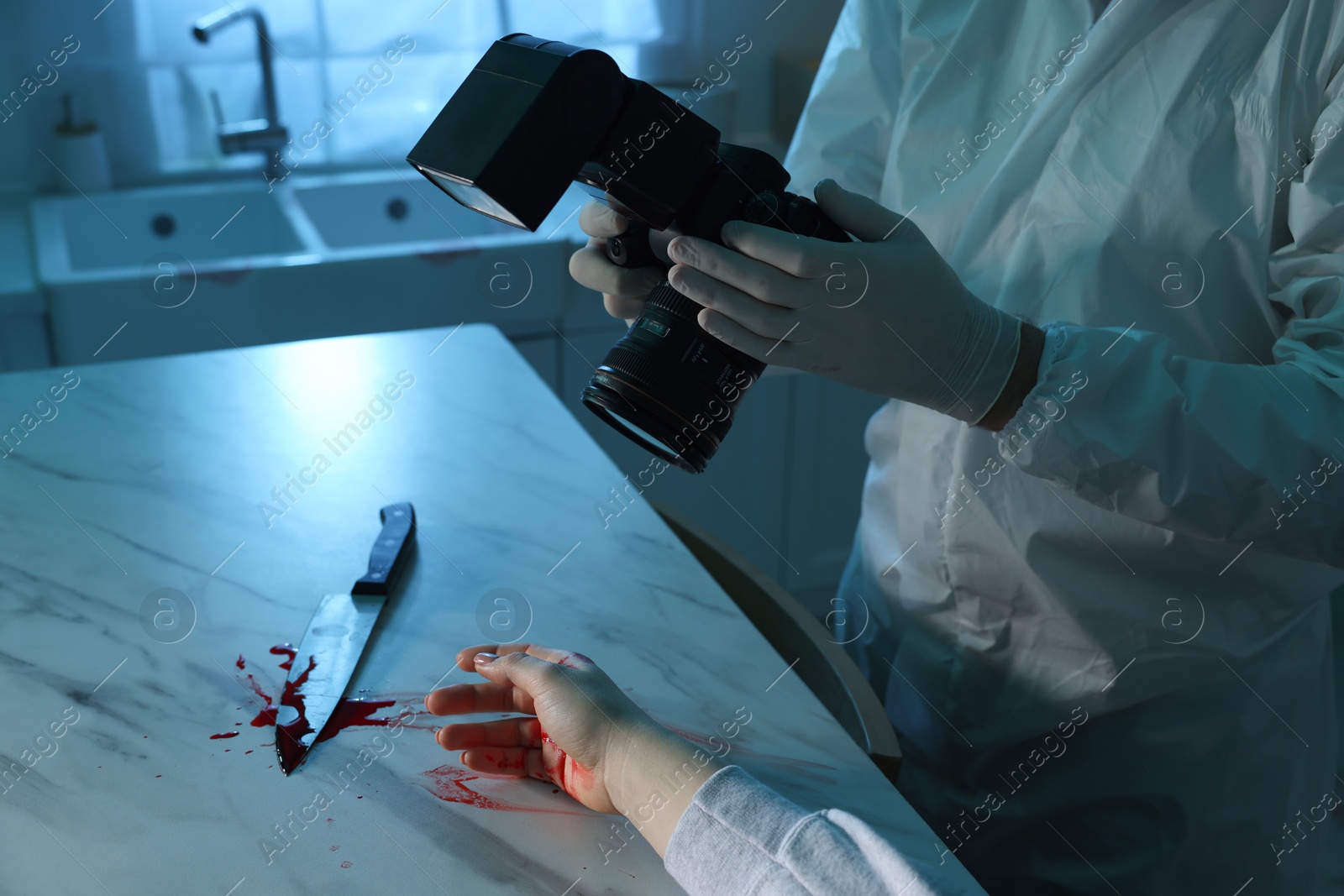 Photo of Forensic expert with camera, knife, blood and dead woman's body at countertop indoors, closeup