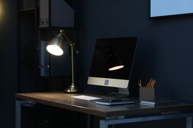 Photo of Comfortable workplace with computer and lamp on wooden desk at home office in evening