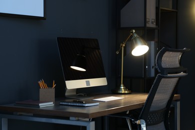 Photo of Comfortable workplace with computer and lamp on wooden desk at home office in evening