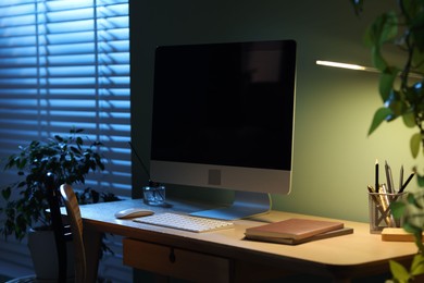 Photo of Comfortable workplace with computer and lamp on wooden desk at home office in evening