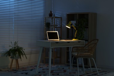 Comfortable workplace with laptop and lamp on desk at home office in evening