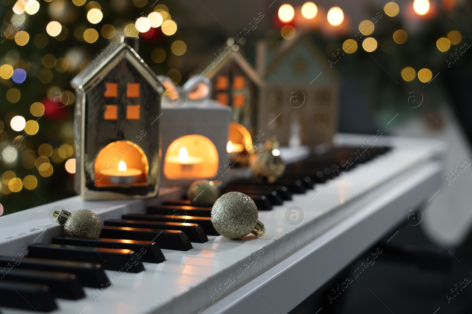 Photo of Christmas ornaments and house shaped holders with burning candles on electric piano against blurred lights, closeup. Bokeh effect