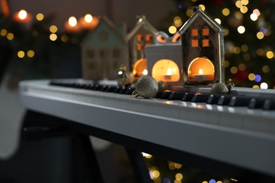 Photo of Christmas ornaments and house shaped holders with burning candles on electric piano against blurred lights, closeup. Bokeh effect