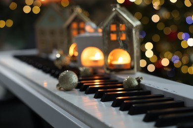 Photo of Christmas ornaments and house shaped holders with burning candles on electric piano against blurred lights, closeup. Bokeh effect