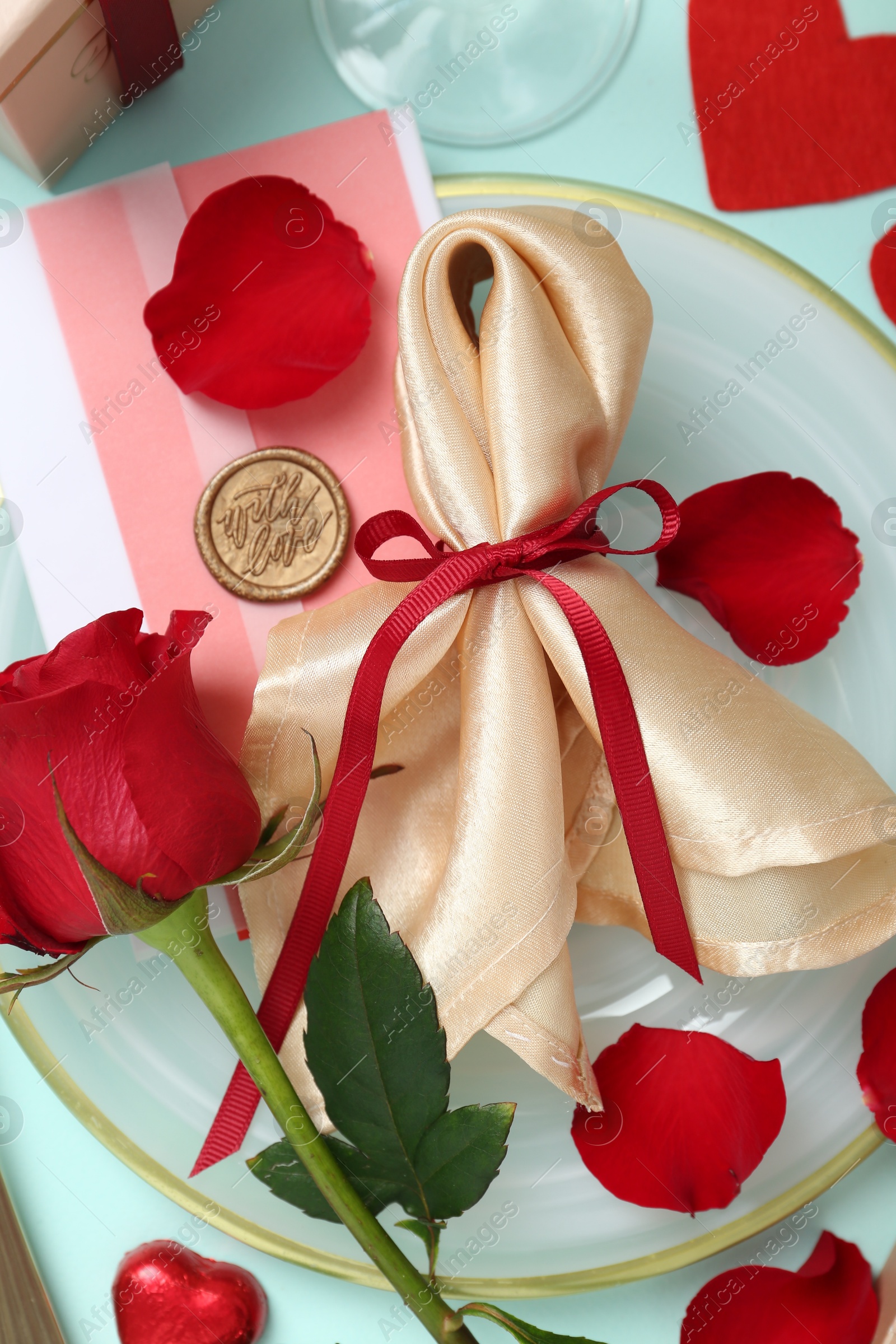 Photo of Romantic place setting with napkin, petals and red rose on light blue background, flat lay. Valentine's day celebration