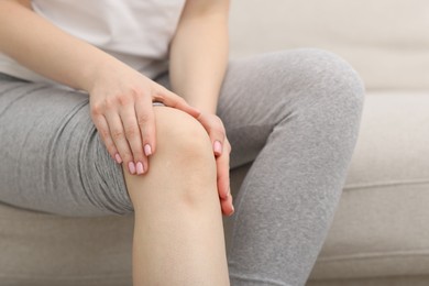 Photo of Young woman suffering from pain in knee on sofa at home, closeup