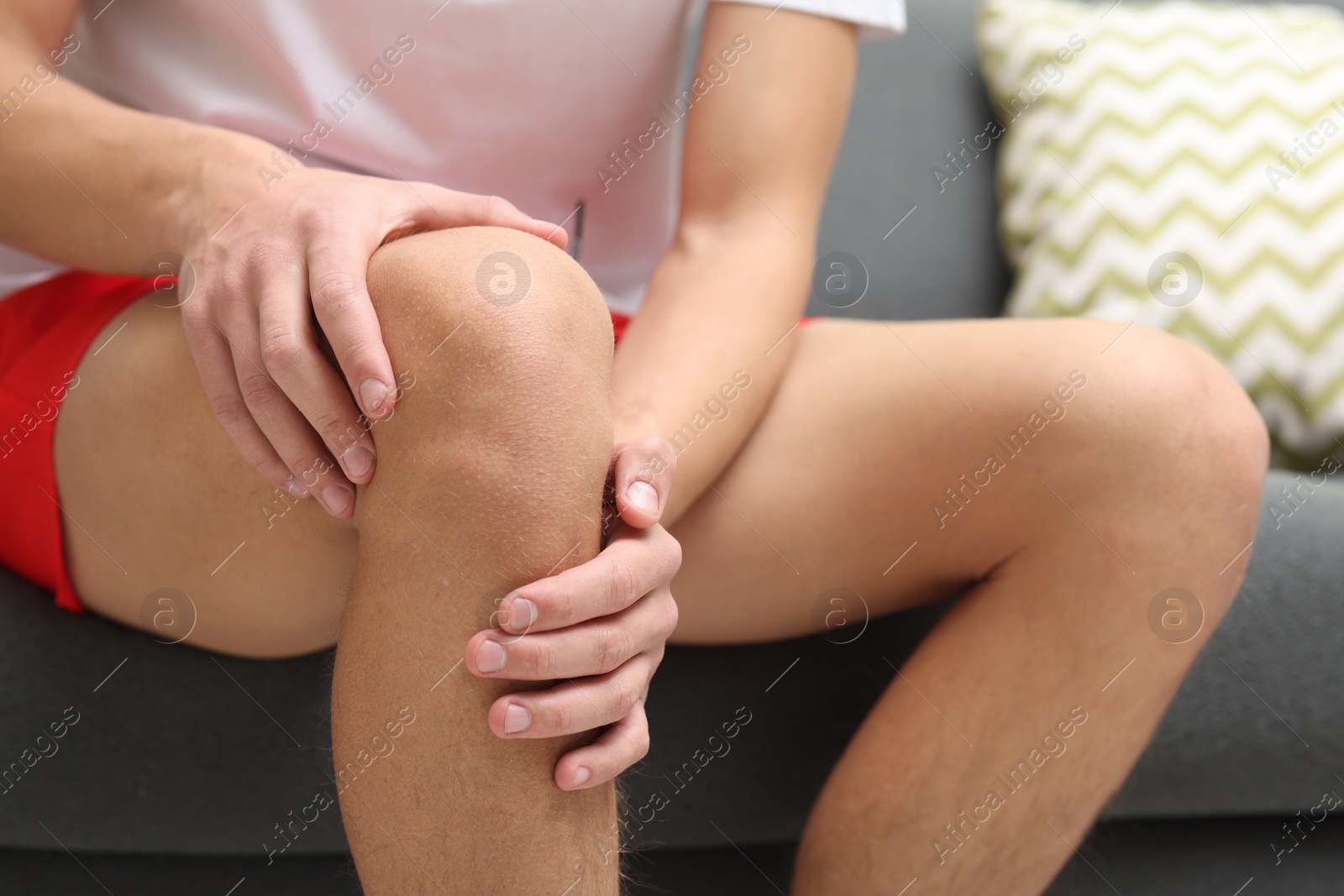 Photo of Young man suffering from pain in knee on sofa at home, closeup