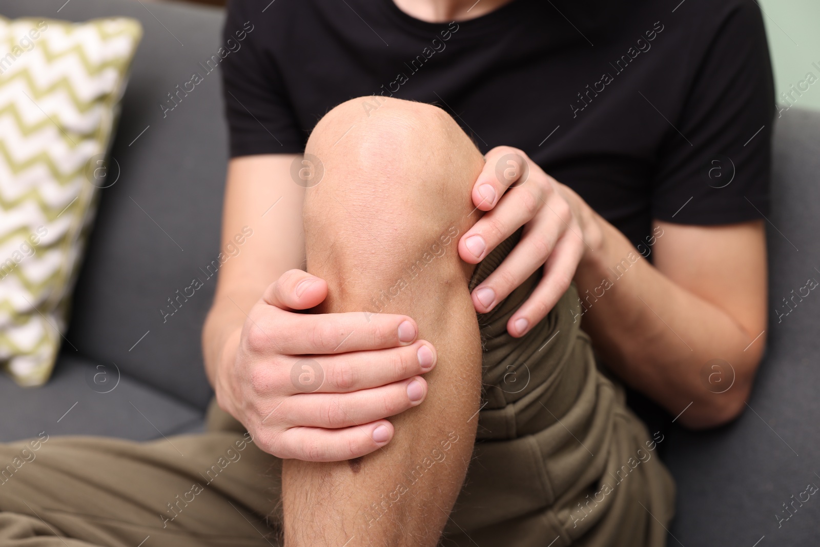 Photo of Young man suffering from pain in knee on sofa at home, closeup